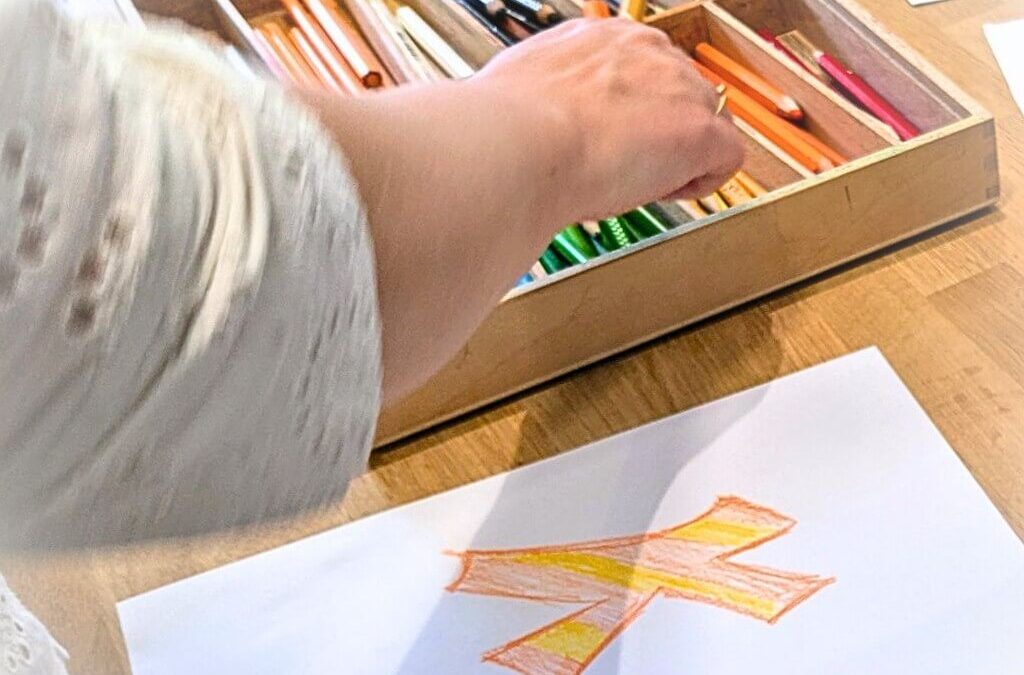 Hånd, farveblyanter, papir med K i gul og orange