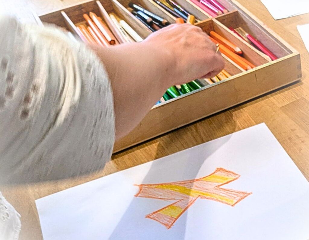 Hånd, farveblyanter, papir med K i gul og orange