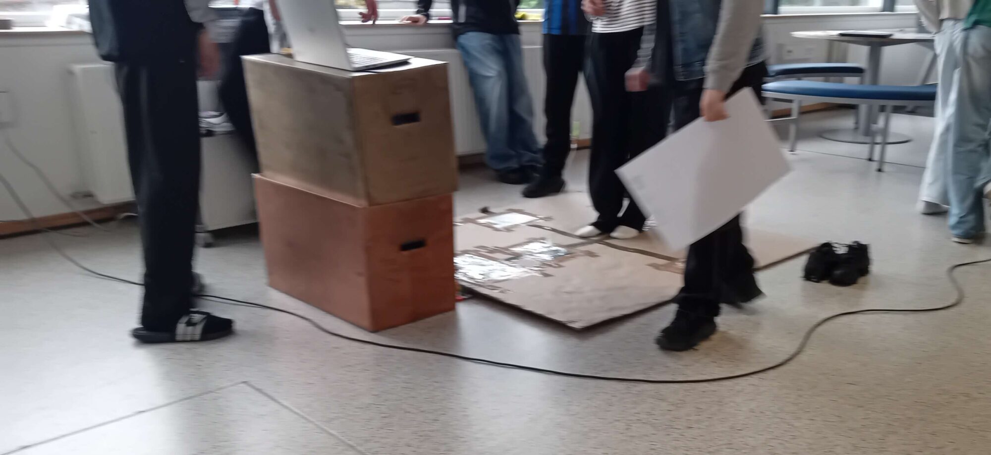 Elever står omkring den hjemmebyggede dancefloor-simulator på gulvet; en af dem holder en planche.